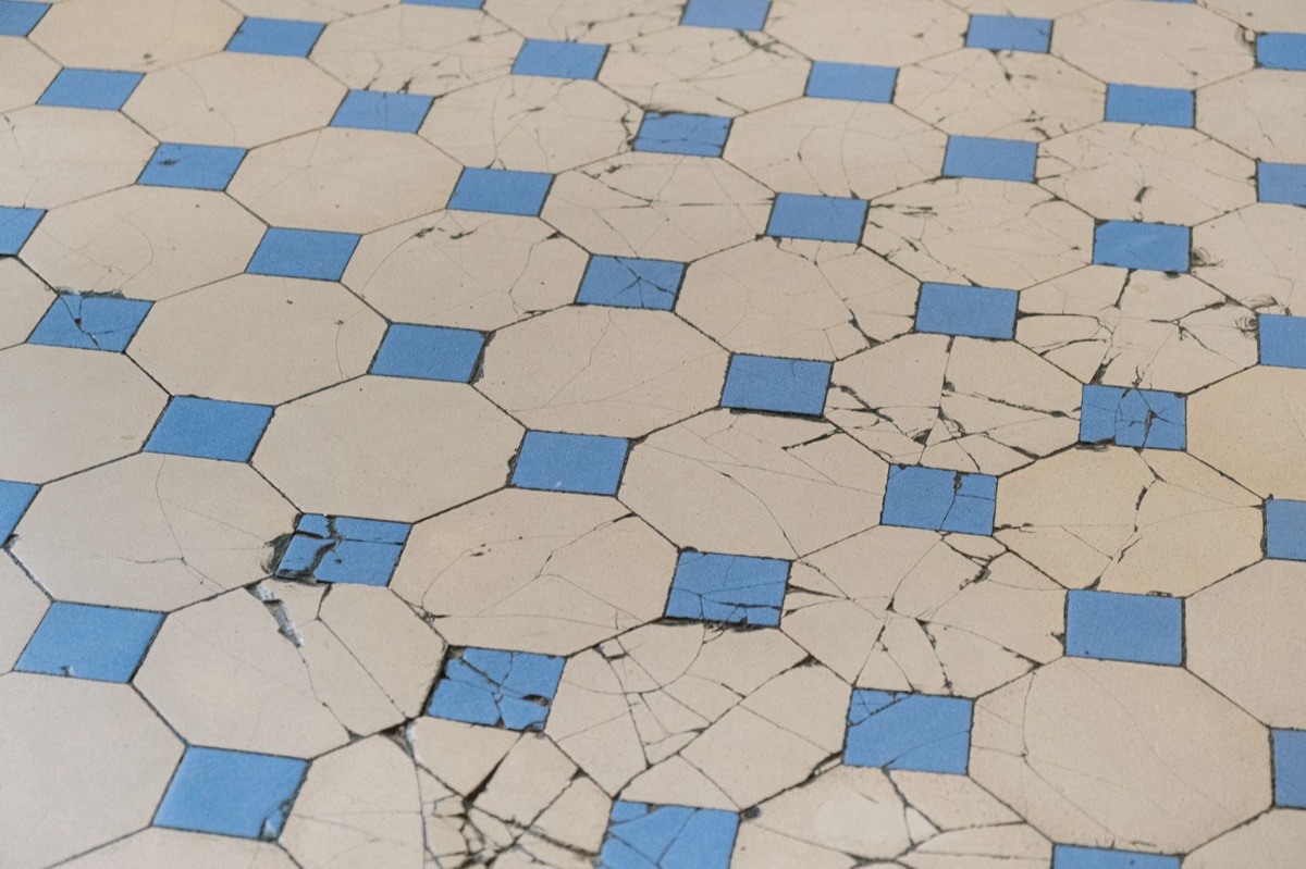 A person in work gloves carefully applies new grout to a section of a tiled floor where old grout was cracked, using a grout float tool.