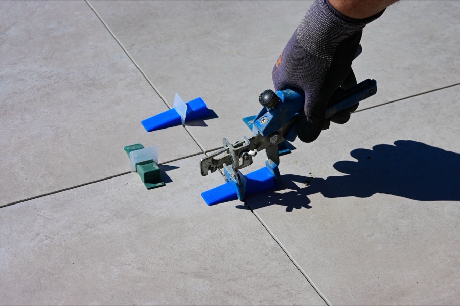 A hand holding a grout saw, carefully scraping out old, cracked grout from between tiles.